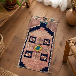 May include: A small, rectangular rug with a faded pink background and a geometric design. The rug features a dark blue border and central design elements, including floral and star-like patterns in various colors. The rug is placed on a wooden floor.