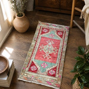 Op de afbeelding: Een rechthoekig vloerkleed in vintage-stijl met een rood middenveld en geometrische patronen in turquoise, roze en wit. Het vloerkleed heeft een versleten uitstraling met een gefranjerde rand, geschikt voor een hal of kleine ruimte. Afmetingen: ongeveer 60 x 120 cm.