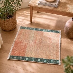 May include: A square, peach-colored rug with a teal border featuring a floral pattern. The rug has a distressed look with a faded appearance. The rug is placed on a wooden floor, with a woven basket, a wooden bench, and a ceramic vase in the background.