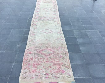 May include: A rectangular, vintage-style rug with a faded, distressed appearance. The rug features a central design of pink and beige geometric patterns, with a border of triangles and fringe. The rug is placed on a dark gray tiled surface.