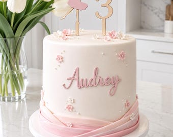 Op de afbeelding: Een cilindrische taart met glad wit glazuur, versierd met kleine roze en witte bloemen en parelaccenten. De naam Audrey staat in roze schrift, met een gouden ballerina en het cijfer 3 als taarttopper. De basis heeft roze stofachtige lagen.