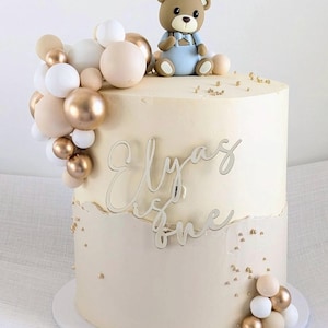 May include: A two-tiered cake with off-white frosting, decorated with a brown teddy bear figurine wearing a blue outfit. The cake is adorned with gold, white, and beige spheres, and the name "Elyaz" and the word "one" are written on the cake.