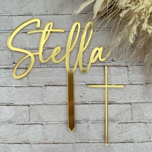 May include: Gold acrylic cake topper with the name "Stella" in a cursive font and a gold cross.