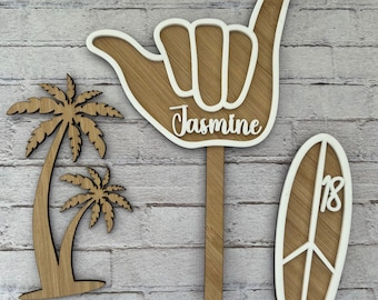 Könnte beinhalten: Drei Holzdekorationen mit Strandthema. Ein Palmenbaum, ein Surfbrett mit der Nummer 18 darauf und eine Hand, die das Shaka-Zeichen macht, mit dem Namen Jasmine darauf.