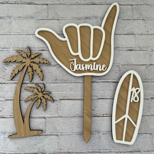 May include: Three wooden decorations with a beach theme. A palm tree, a surfboard with the number 18 on it, and a hand making the shaka sign with the name Jasmine written on it.