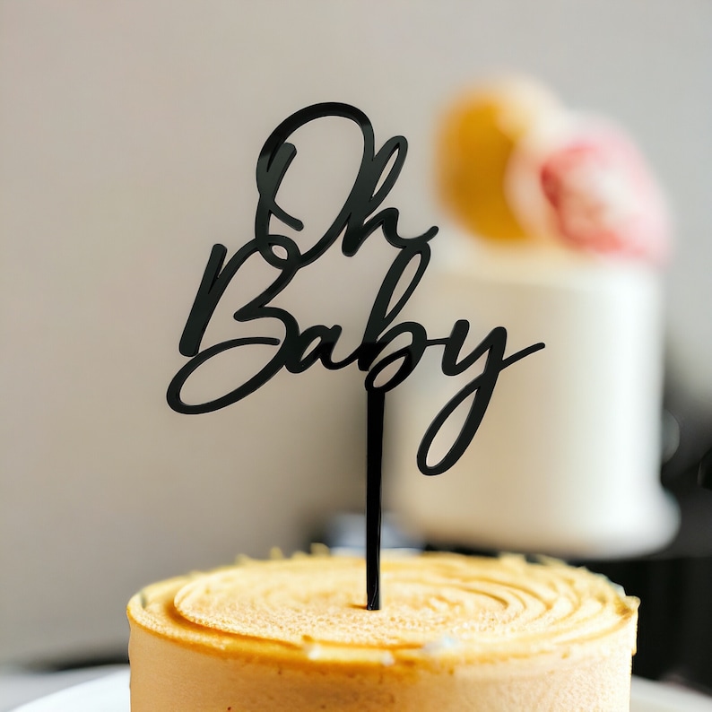 May include: Black acrylic cake topper with the text "Oh Baby" in a cursive font. The topper is on top of a round, light brown cake.