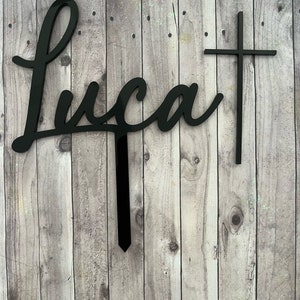 May include: Black acrylic cake topper with the name "Luca" written in cursive and a cross to the right.