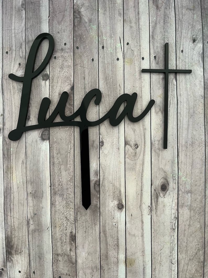 May include: Black acrylic cake topper with the name "Luca" written in cursive and a cross to the right.