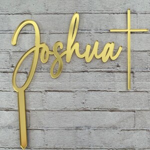 May include: Gold-colored acrylic cake topper with the name "Joshua" in cursive script and a cross.