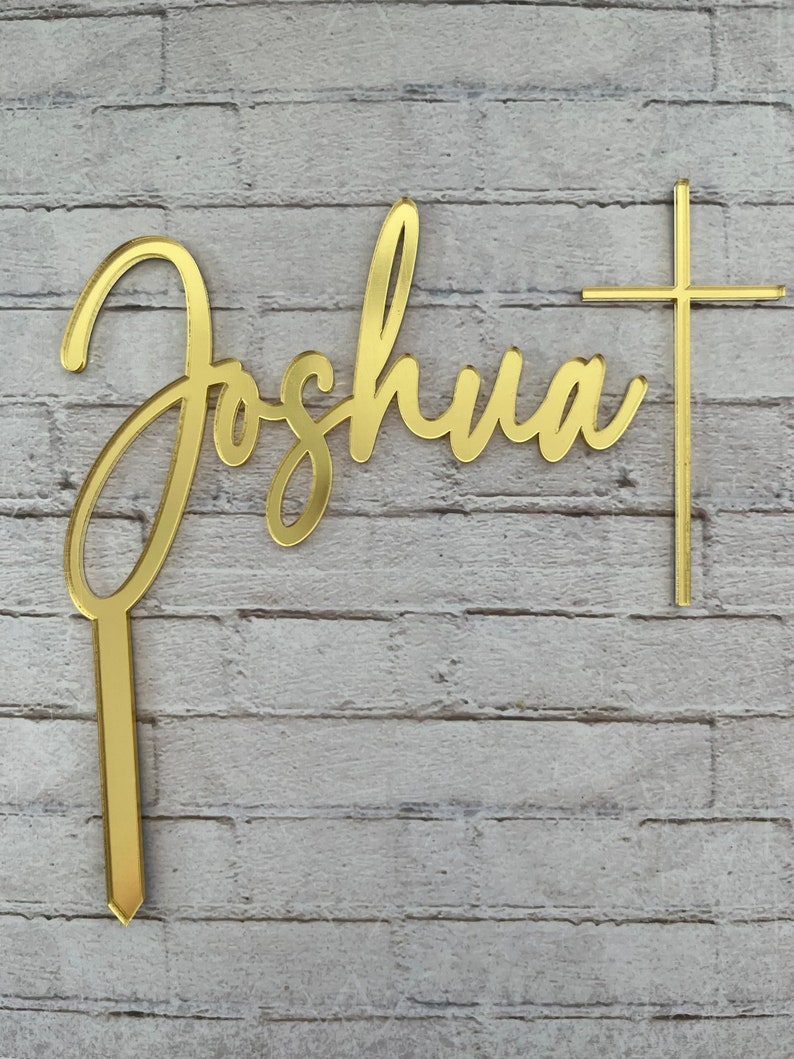 May include: Gold-colored acrylic cake topper with the name "Joshua" in cursive script and a cross.