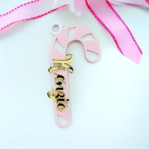 May include: A pink and white candy cane-shaped ornament with the name "Kenzie" in gold script. The ornament has a small hole at the top for hanging and is set against a white background with pink ribbon.