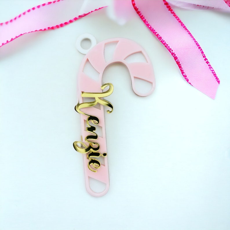 May include: A pink and white candy cane-shaped ornament with the name "Kenzie" in gold script. The ornament has a small hole at the top for hanging and is set against a white background with pink ribbon.