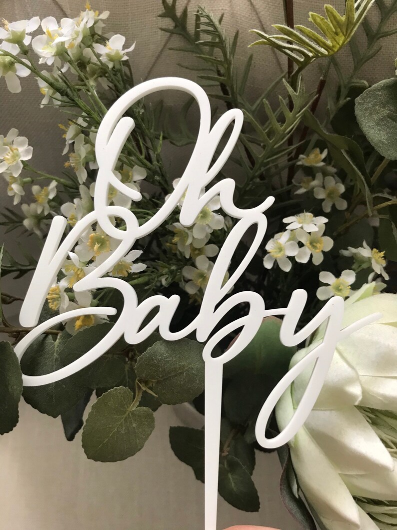 May include: White acrylic cake topper featuring the words "Oh Baby" in a cursive script. The topper is surrounded by green foliage and white flowers.