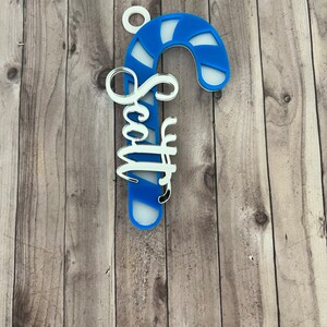 May include: A blue and white candy cane ornament with the name "Scott" in a cursive font. The ornament has a white loop at the top for hanging. The background is a wooden surface.