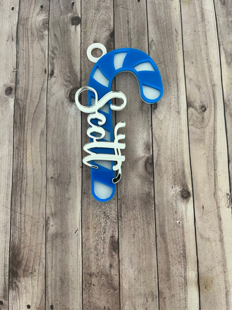May include: A blue and white candy cane ornament with the name "Scott" in a cursive font. The ornament has a white loop at the top for hanging. The background is a wooden surface.