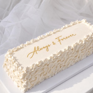 May include: A rectangular cake with smooth off-white frosting on top and ruffled frosting along the sides. The text "Always & Forever" is written in gold cursive script on the top surface of the cake. The cake sits on a white rectangular base.