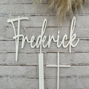May include: White wooden name sign with the name "Frederick" in cursive script. The sign is accompanied by two white wooden stakes, one shaped like a cross and the other a simple vertical stake.