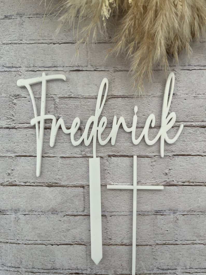 May include: White wooden name sign with the name "Frederick" in cursive script. The sign is accompanied by two white wooden stakes, one shaped like a cross and the other a simple vertical stake.