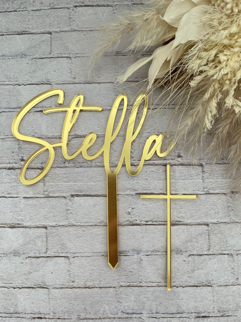 May include: Gold acrylic cake topper with the name "Stella" in a cursive font and a gold cross.