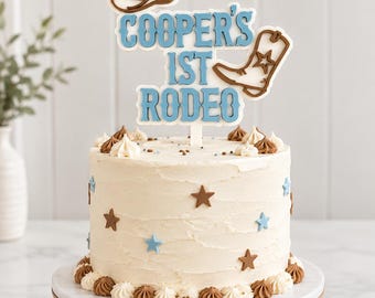 May include: A round cake with white frosting, decorated with small brown and light blue stars and piped frosting swirls at the base. A cake topper features the text COOPER'S 1ST RODEO in light blue, with a brown cowboy hat and boot icon.