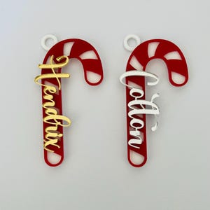 May include: Two candy cane ornaments with personalized names. The candy canes are red with white stripes and a white loop for hanging. One has a gold name, the other a silver name. Festive holiday decor.