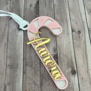 May include: A pink and white candy cane-shaped Christmas ornament with the name "Aurora" in gold script. The ornament has a white ribbon for hanging. The design is a festive holiday decoration.