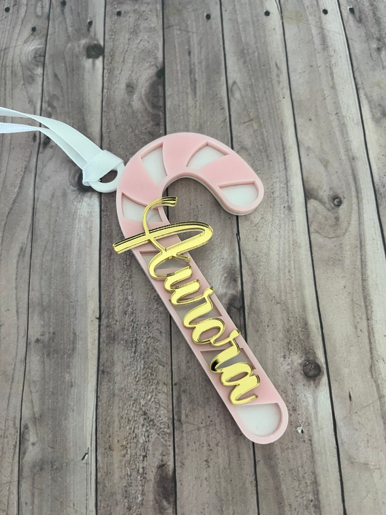 May include: A pink and white candy cane-shaped Christmas ornament with the name "Aurora" in gold script. The ornament has a white ribbon for hanging. The design is a festive holiday decoration.