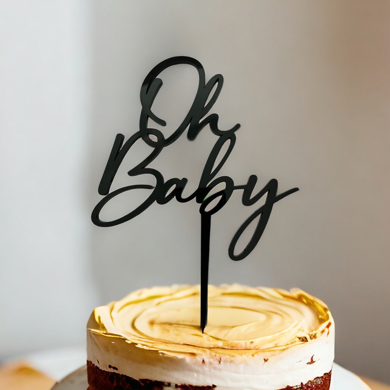 May include: Black acrylic cake topper with the words "Oh Baby" in a cursive font. The topper is on top of a frosted cake.