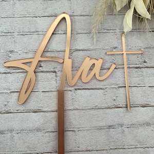 May include: A gold-colored cake topper with the word "Ava" in a cursive script and a small cross. The topper is designed to be inserted into a cake.