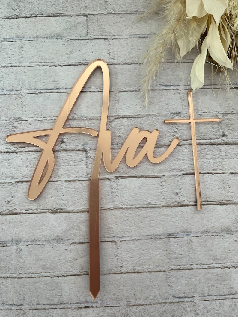 May include: A gold-colored cake topper with the word "Ava" in a cursive script and a small cross. The topper is designed to be inserted into a cake.