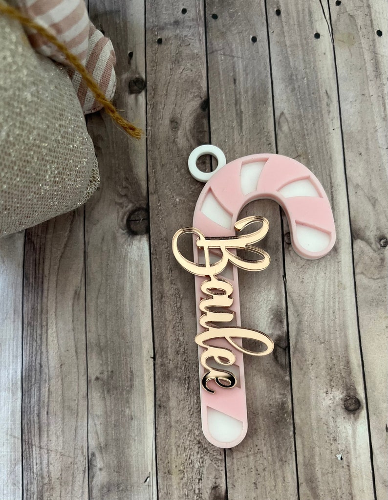 May include: A pink and white candy cane Christmas ornament with the name "Bailee" in gold script. The ornament has a white loop at the top for hanging. The background is a wooden surface.