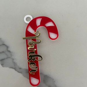 May include: A red and white striped candy cane Christmas ornament with the name "Parker" in gold script. The ornament has a white loop at the top for hanging.