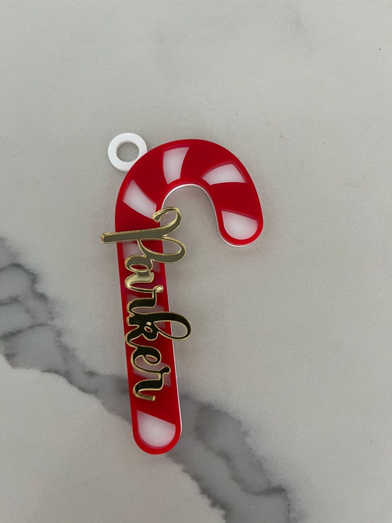 May include: A red and white striped candy cane Christmas ornament with the name "Parker" in gold script. The ornament has a white loop at the top for hanging.