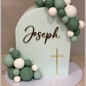 May include: A light green cake decorated with a gold cross and the name Joseph. The cake is surrounded by green and white balloons.