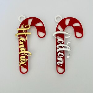 May include: Two candy cane ornaments with personalized names. The candy canes are red with white stripes and a white loop for hanging. One has a gold name, the other a silver name. Festive holiday decor.