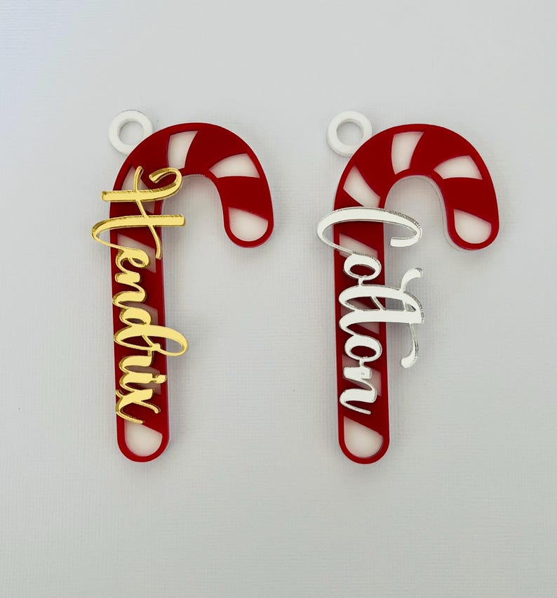 May include: Two candy cane ornaments with personalized names. The candy canes are red with white stripes and a white loop for hanging. One has a gold name, the other a silver name. Festive holiday decor.
