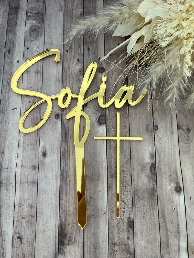 May include: Gold acrylic cake topper with the name "Sofia" in cursive script and a gold cross.