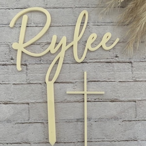 May include: Wooden cake topper with the name "Rylee" in cursive script and a simple cross. The topper is light brown and is perfect for a baptism or christening cake.