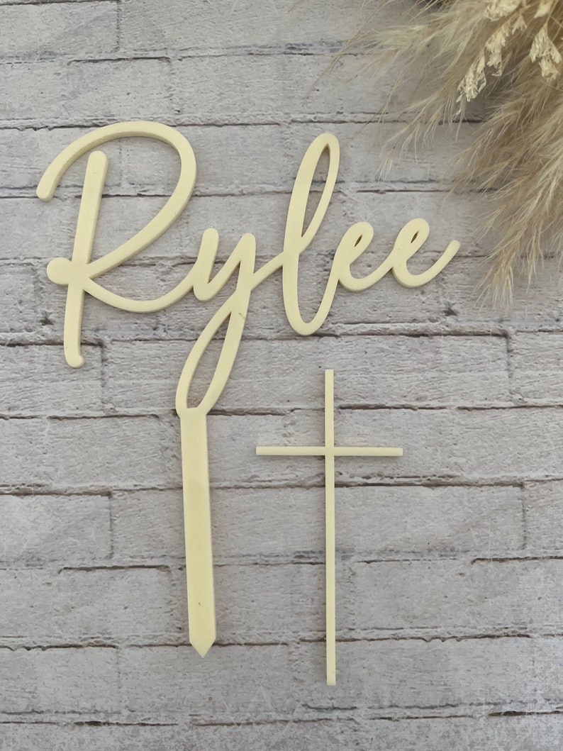 May include: Wooden cake topper with the name "Rylee" in cursive script and a simple cross. The topper is light brown and is perfect for a baptism or christening cake.