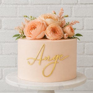 May include: A round, light peach-coloured cake topped with a cluster of soft peach-coloured flowers and green leaves. The front of the cake features a gold-coloured script topper that reads "Ange" in a cursive font, centred on the smooth surface.