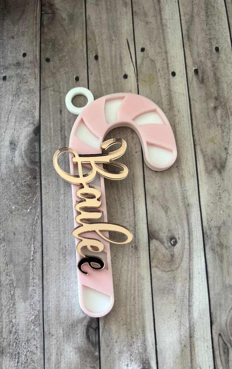 May include: A pink and white candy cane ornament with the name "Barbie" in gold script. The ornament has a white loop at the top for hanging. The candy cane design is a festive holiday decoration.