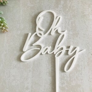 May include: White acrylic cake topper with the words "Oh Baby" in a cursive font.