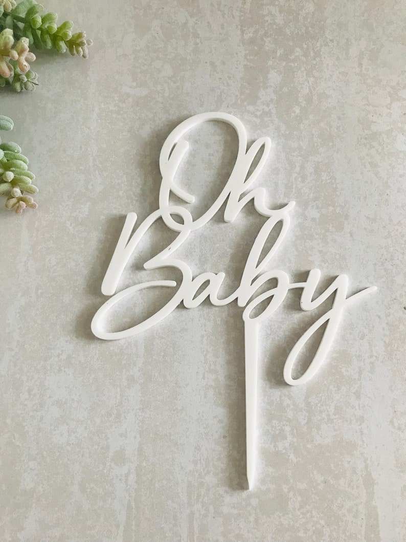 May include: White acrylic cake topper with the words "Oh Baby" in a cursive font.
