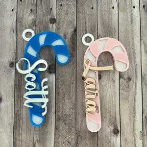 May include: Two candy cane ornaments, one blue and one pink, with white accents. The blue candy cane has the name "Scott" in silver script, and the pink one has "Laura" in gold script. Both have a small white loop for hanging.