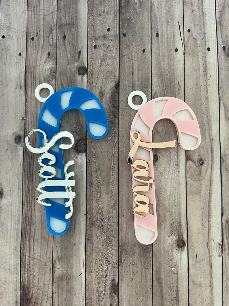 May include: Two candy cane ornaments, one blue and one pink, with white accents. The blue candy cane has the name "Scott" in silver script, and the pink one has "Laura" in gold script. Both have a small white loop for hanging.