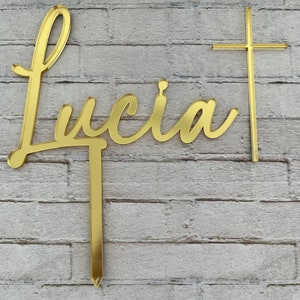 May include: Gold-colored acrylic cake topper with the name "Lucia" in a cursive script and a cross. The topper is designed for a cake.