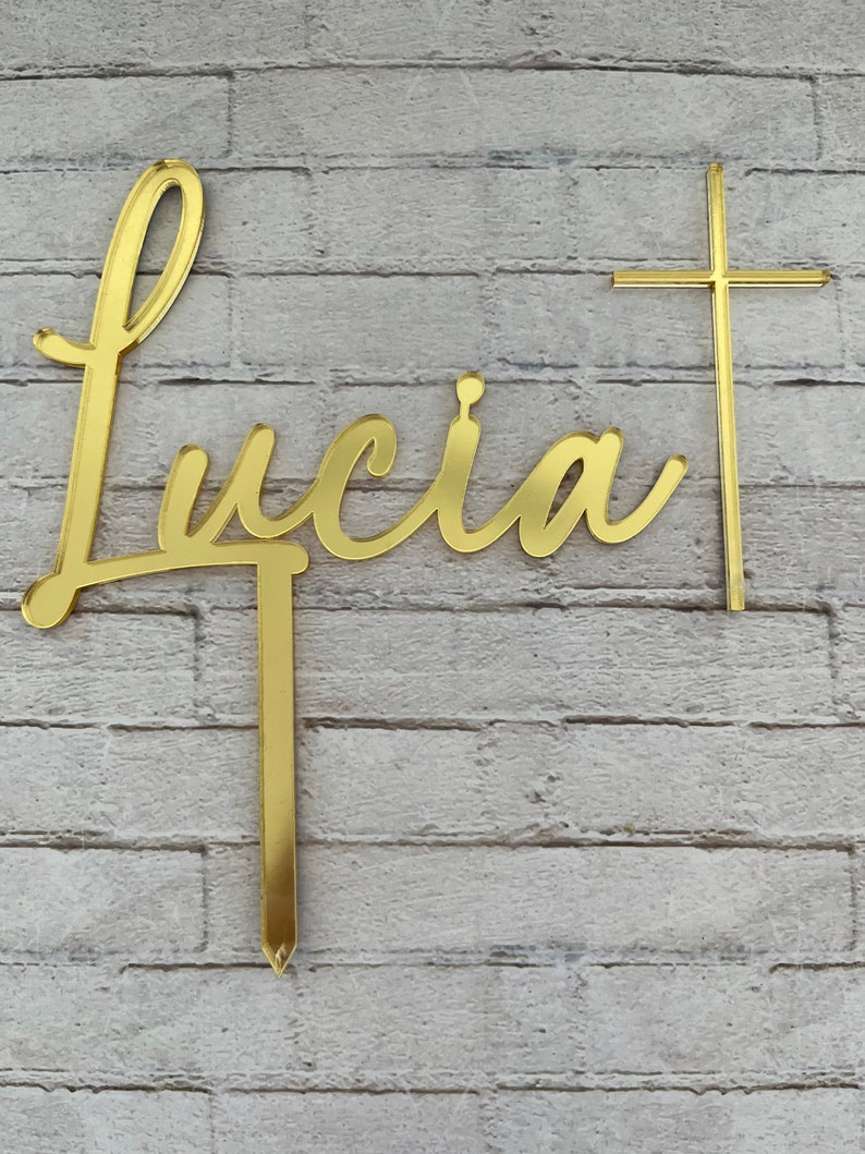 May include: Gold-colored acrylic cake topper with the name "Lucia" in a cursive script and a cross. The topper is designed for a cake.