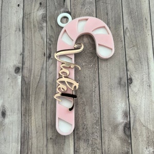 May include: A pink and white candy cane ornament with the word "Tia" in gold script. The ornament has a white loop at the top for hanging. The candy cane design is a festive holiday decoration.