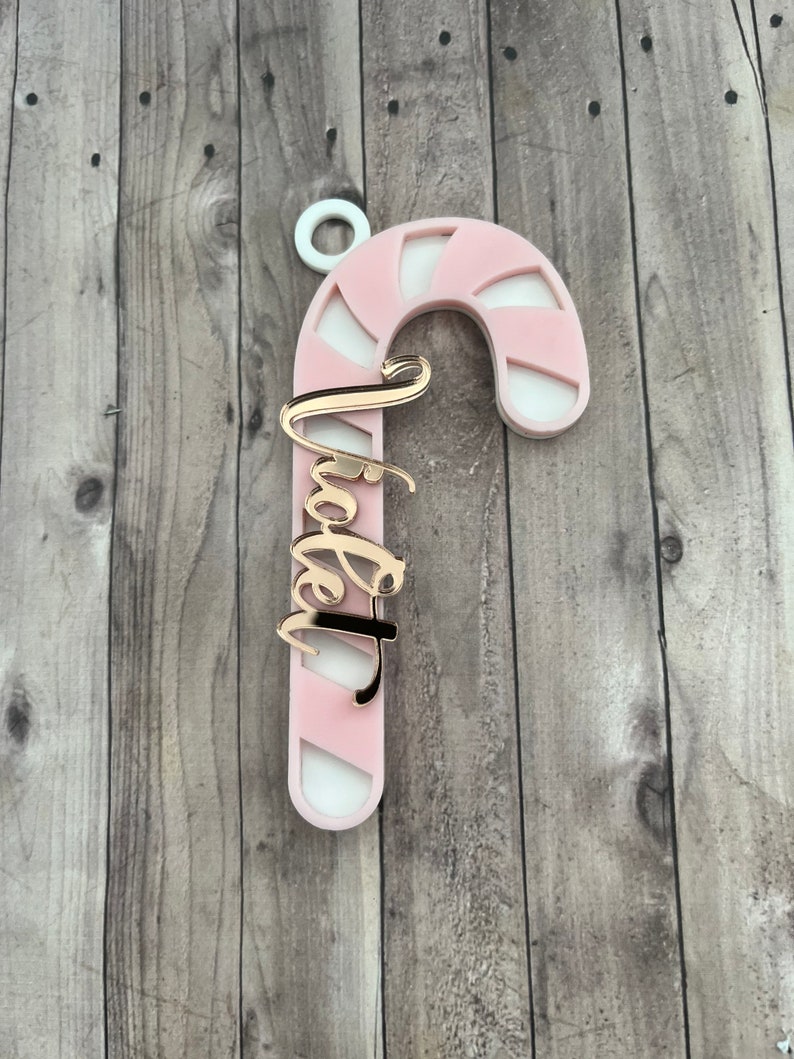 May include: A pink and white candy cane ornament with the word "Tia" in gold script. The ornament has a white loop at the top for hanging. The candy cane design is a festive holiday decoration.