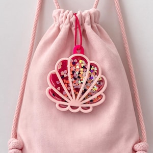 May include: A pale pink drawstring bag with a seashell-shaped charm. The charm is pink with a clear centre filled with heart-shaped confetti. The drawstring is pink.